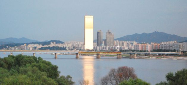 View of Yeouido island's iconic 63 building, lit up in bright sunshine, reflected in the Hangang river, with the cityscape and mountains behind.