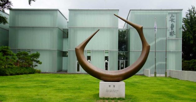 Exterior view of the Ahn Jung-geun Memorial Hall, comprised of three glass buildings, in front of them a brown sickle statue on a bright green lawn.