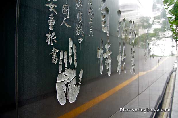 A black polished stone wall, with a hand print etched into it, and Korean lettering around, with a yellow stripe running underneath.