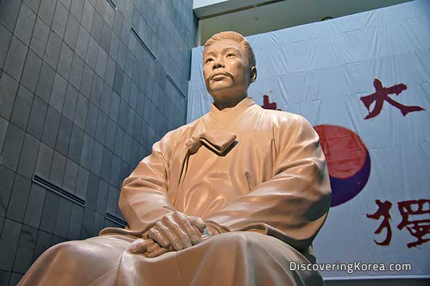 A statue of Ahn Jung in light stone, on a backdrop of a stone wall and a fabric hanging with a red and blue yin-yang sign and Korean lettering.