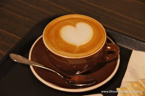 Close up of a brown cup with cappuccino, with a heart shape on the top, a spoon on the saucer on a wooden surface.