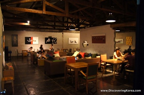 Dimly lit interior of Anthracite Cafe in Seoul, wooden chairs and tables in the foreground, in the background some low seating and white walls with modern artwork.