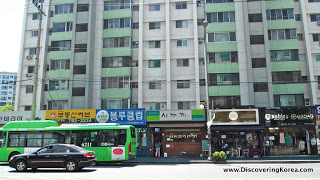 A grey and green high rise apartment block in Yongsan, with shopfronts at street level, and traffic on the road in front.