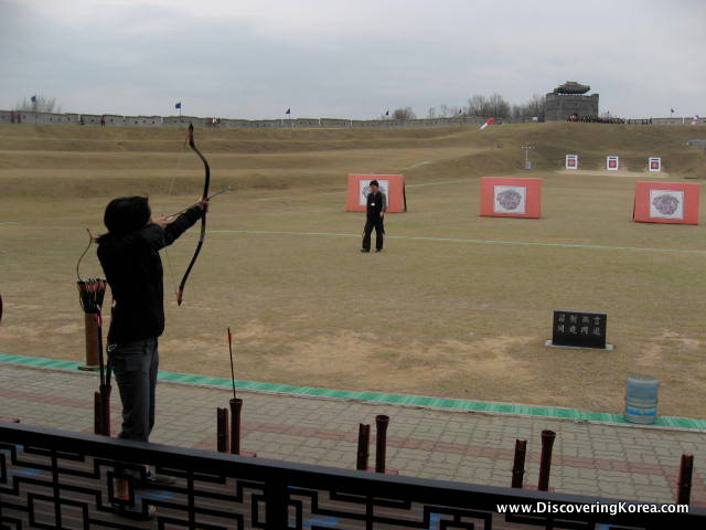 A woman with an archery bow, and six targets in the background, on grass.