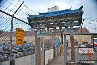 A gate flanked in wire fencing and barbed wire. An ornate roof over the small entranceway with words "Camp Kim" printed above.