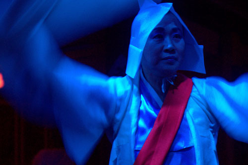 Close up of a woman with traditional robes and headwear, with a red sash dancing, in a blue light on a black background.