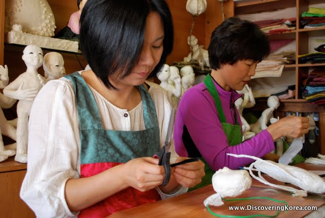 Two ladies at Jeonju hanok village sewing in a fabric shop with dolls in the background.