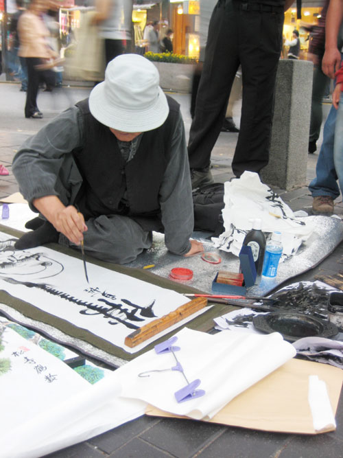 A man sits on the sidewalk, painting a picture on a white paper, with various art supplies around him.