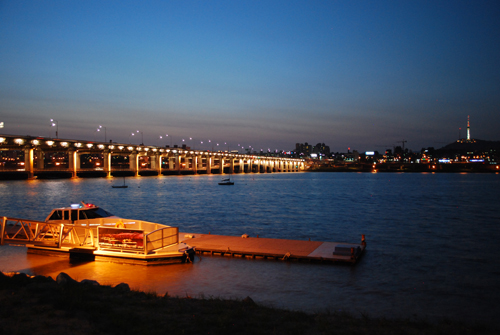 Night view of Hangang river, with the bridge to the left of the frame and a lit up boat at a jetty in the foreground. The city lights are visible in the background.