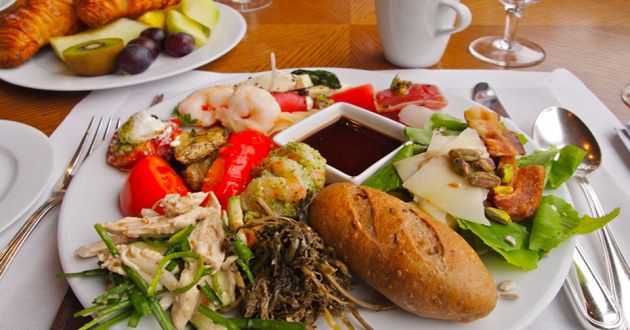 A white plate with various different food, a bread roll, some shrimp, some chicken and soy sauce in a smaller bowl in the center, in the background is a plate with croissant and fruit, on a white place mat on a wooden table.