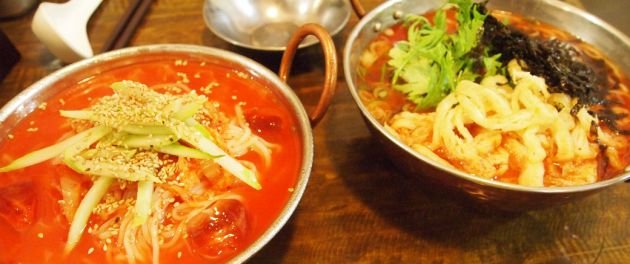 Two metal bowls, one containing noodles in a red broth, and the other with noodles, seaweed and greens, on a wooden table.