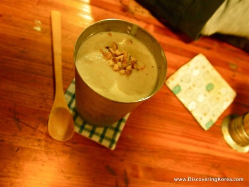 Close up of a metal cup containing a milky drink with nuts on the top, on a wooden table with a wooden spoon next to it.