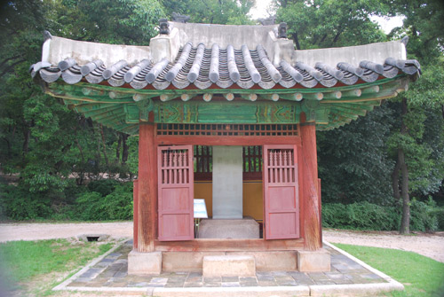 A small wooden house in deep red, with turquoise eaves and a traditional curved roof, surrounded by a stone plinth. Through the open door a concrete block. Pine forest in the background.