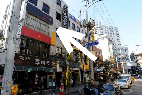 A street of shops and restaurants, with cars to the right of the frame, and a large white arrow pointing to a first floor location.