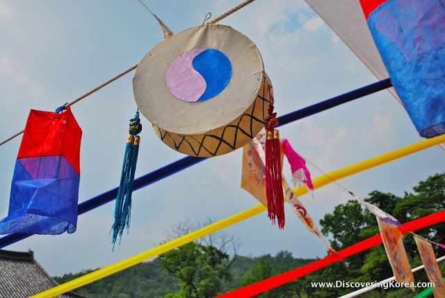 A large drum, with a pink and blue yin-yang symbol hangs from a line, with paper lights in blue and red. In the background is blue sky with clouds and trees.
