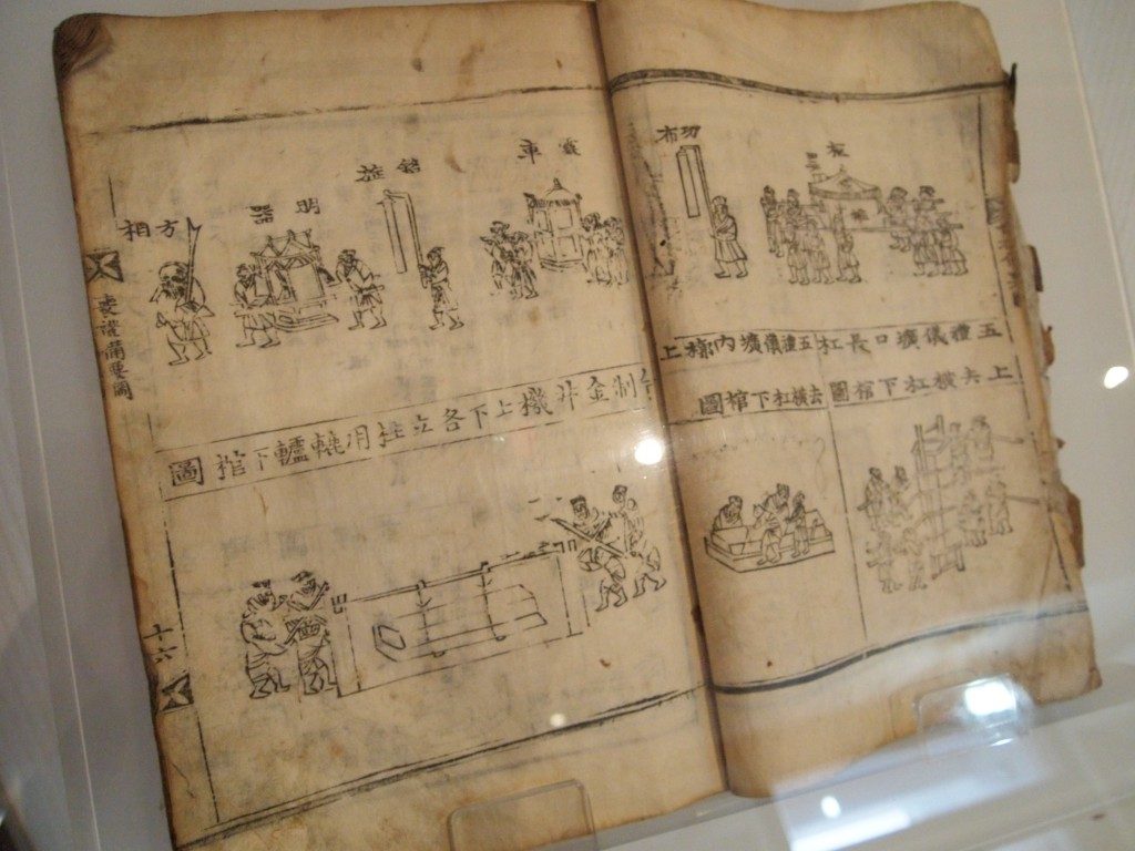 An open book of prayers, with Korean lettering and pen and ink drawn images, on a white background, behind a glass screen at Musee Shuim.
