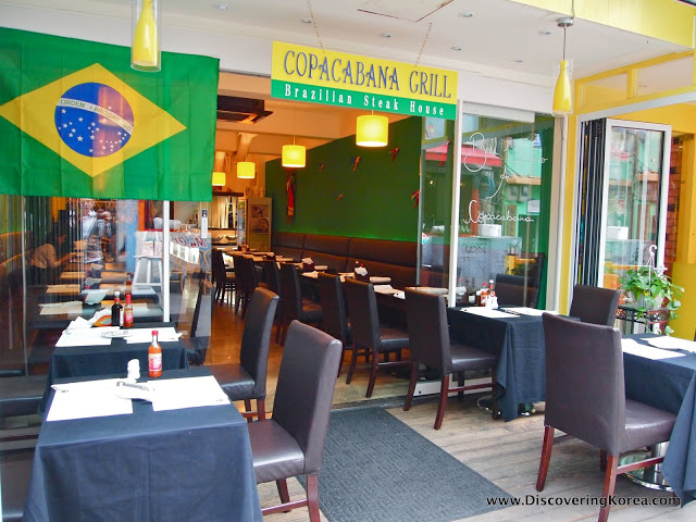 Looking into a Brazilian restaurant, with outside tables, dark brown chairs, dark blue tablecloths. Through the glass doors is more seating, with green walls and yellow lighting.