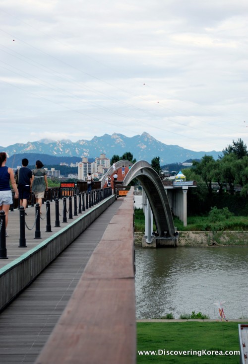 View across Rainbow Bridge, Seonyudo, showing pedestrians on the left, walking across the bridge, the river to the right of the frame and city and mountains in the background.