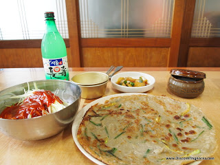 A table set with a dish of buckwheat pancakes, a noodle dish with a red sauce in a metal bowl, condiments behind and bottle of rice wine.