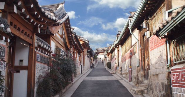 A narrow street in Bukchon Hanok Village with wooden buildings on either side, decorated with red and white tile work. Blue sky and clouds in the background.