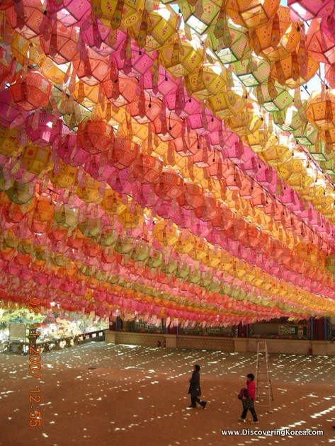 Paper lights at the top of the frame in pink, orange, light-green and yellow above an open area with two people walking.