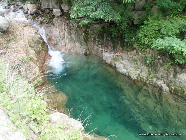 A waterfall from the right of the frame empties into a crystal clear pool of water in a gully, cut out of the rocks in Bukhansan national park.