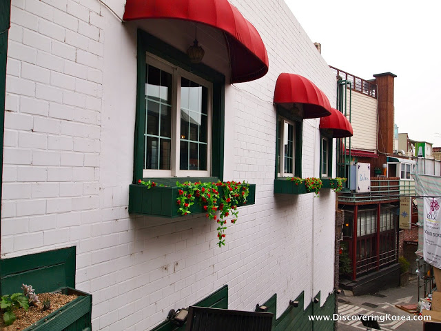 The side of a white brick building, with green painted windows, and trailing plants from the windowboxes. Above the windows are red shade overhangs, at Seoul's only Bulgarian restaurant.