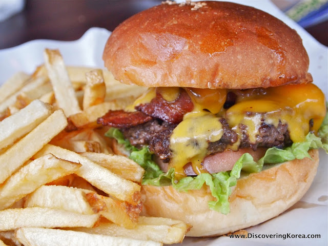 A close up of a burger with bacon and melting cheese, on a white paper background, with fries on the left of the frame.