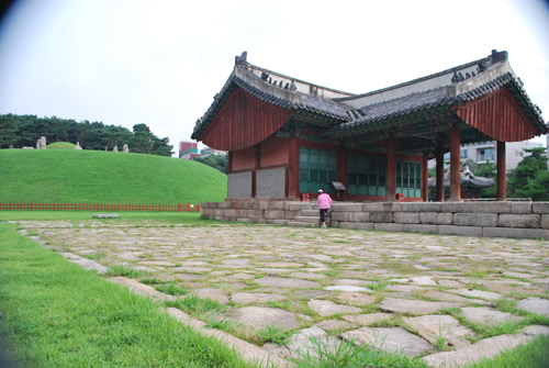 A wooden building with a traditional curved roof, with a stone courtyard surrounded by grass, the burial site of King Seongjong.