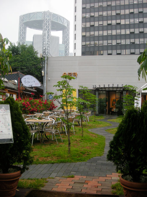 The outside seating area of a cafe, with chairs and tables to the left of the frame, a sunflower and a paved walkway into a large building, with a metal structure in the background.