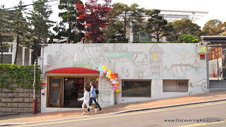 Two people walking downhill on a street, outside a quirky cafe, with murals on the white walls. In the background are trees.