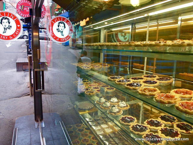 A cafe display cabinet showing various different cakes and tarts, on glass shelves. To the right of the frame is the cafe door leading to the street.