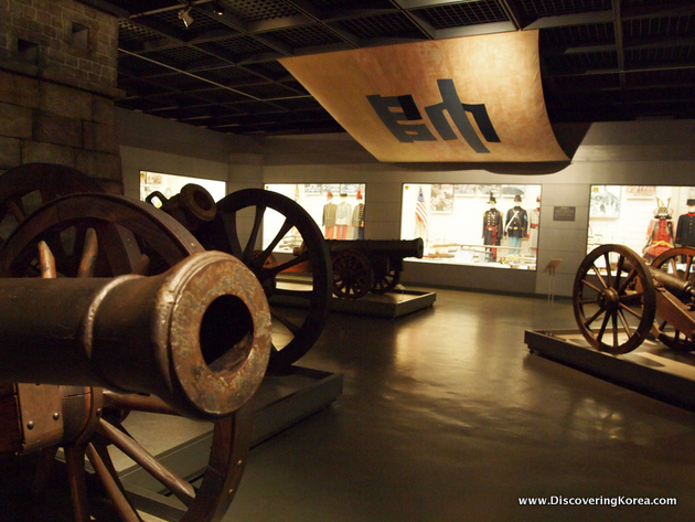 Cannons on a concrete surface inside the Seoul War Memorial. Hanging from the ceiling is fabric printed in Korean. In the background are glass cases containing other displays.