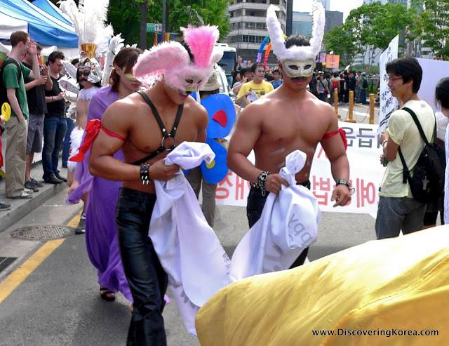 Two men dressed in leather trousers, one wearing a pink feathered face mask and the other wearing a white feathered face mask, walk through a crowd of people carrying a piece of white cloth.