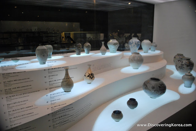 A glass display case with ceramic pottery, some small vases, others a little larger, all set on white surface, spotlit with a dimly lit background.