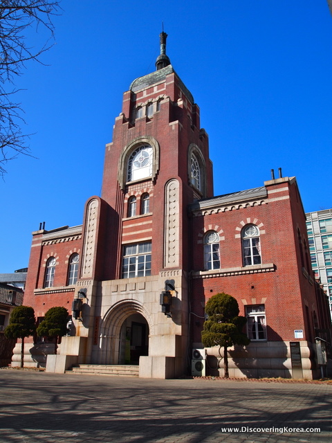 The red brick exterior of Cheondogyo Temple Seoul, a tall tower in the center, an arched stone entrance with stone steps leading up, blue sky in the background.