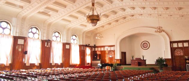 Inside the Cheondogyo Temple, rows of seats facing an altar, lights hanging from the white ceiling, with sun shining through the curtains on the arched windows.