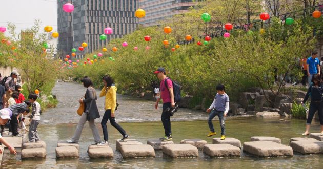 Paving stones crossing the Cheonggyecheon stream with people walking and multicolored paper lanterns in the background.