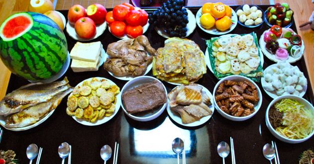 A black table, laden with plates and bowls of different food. A large green watermelon on the left of the frame, with whole fish, various meats and fruits and noodle dishes to the right of the frame.