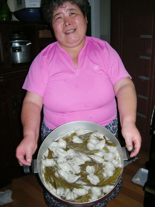 A Korean lady in a pink shirt holds out a metal saucepan full of white dumplings, a traditional meal at Chuseok.