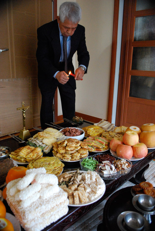 A man lights a candle over a table full of food for Chuseok. Plates of various Korean specialties on a black surface.