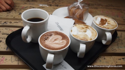 Four cups of coffee on a black tray, on a wooden table with a white plate and a packet of cookies.