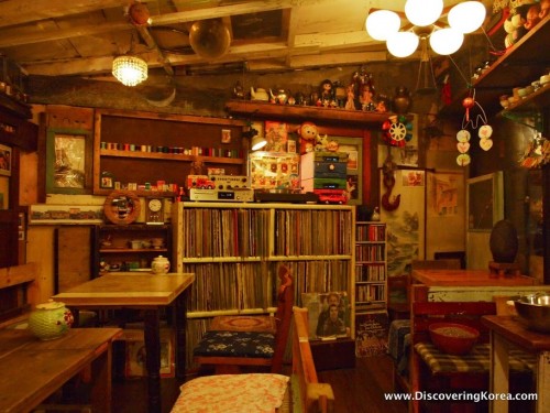 A dimly lit room at Coffee Han cafe in Seoul, with wooden tables and chairs, a bookcase of old LP records, and various curios and ornaments on wooden shelves.