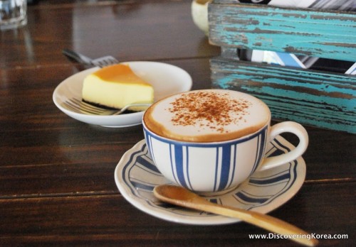 A blue and white striped cup with cappuccino coffee, wooden spoon on the saucer, with a cake on a white plate in the background, on a wooden surface.