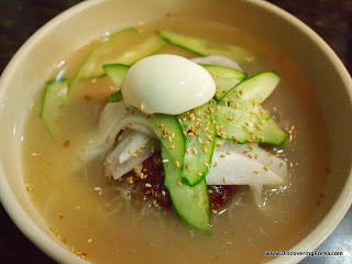 A clear broth noodle soup, with green vegetables and an half an egg on the top, in a light brown ceramic bowl.