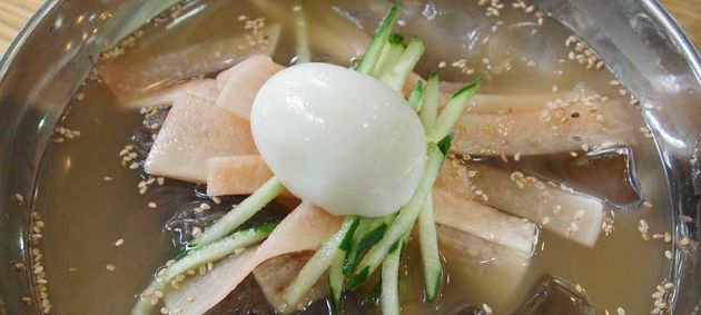 Close up of a metal bowl with a light broth, containing noodles, thinly sliced cucumber garnish and a boiled egg on top.