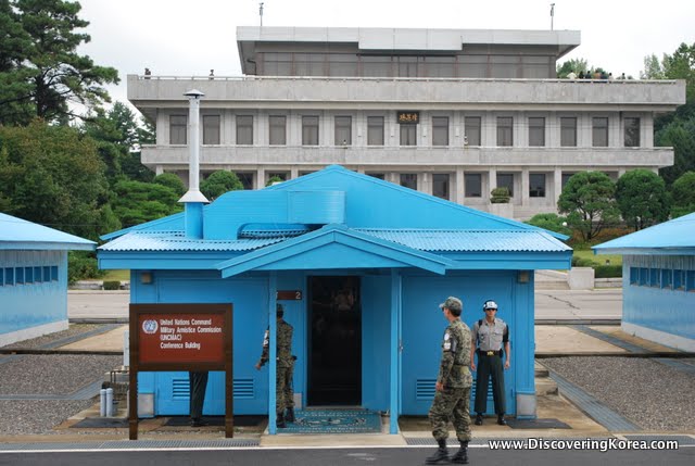Entrance to DMZ at Panmunjeom, a blue building flanked by a large stone building in the background. Three soldiers outside the blue building and a brown sign to the left of the frame.