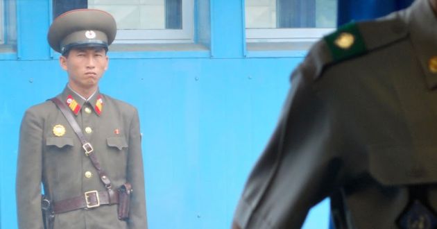 A Korean soldier stands against a blue wall, with a soft focus close up of another soldier's shoulder in the foreground.