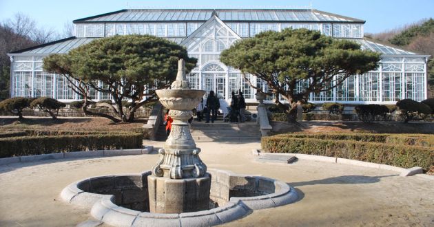 Exterior view of Daeonsil, a large glass house with white framework, an A frame entrance. In front is a courtyard with a stone fountain leading up to the steps, two large trees flanking the entrance.