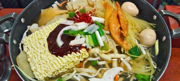 Close up of a metal pan containing the ingredients for ddeokbokki, noodles, chopped vegetables, eggs and meat.